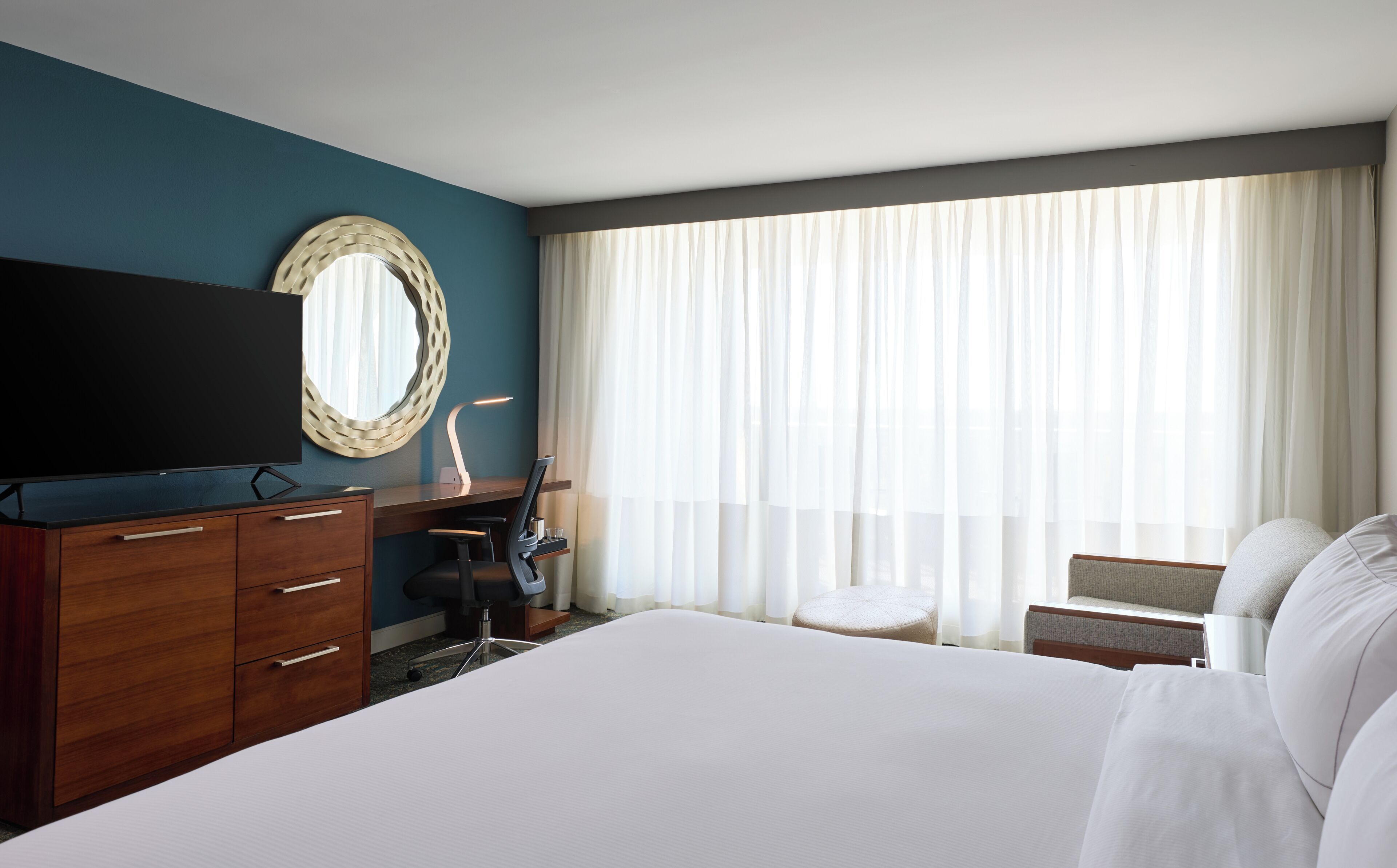 Een hotelkamer met een bed, een bureau met stoel, een televisie en een groot raam met gordijnen.