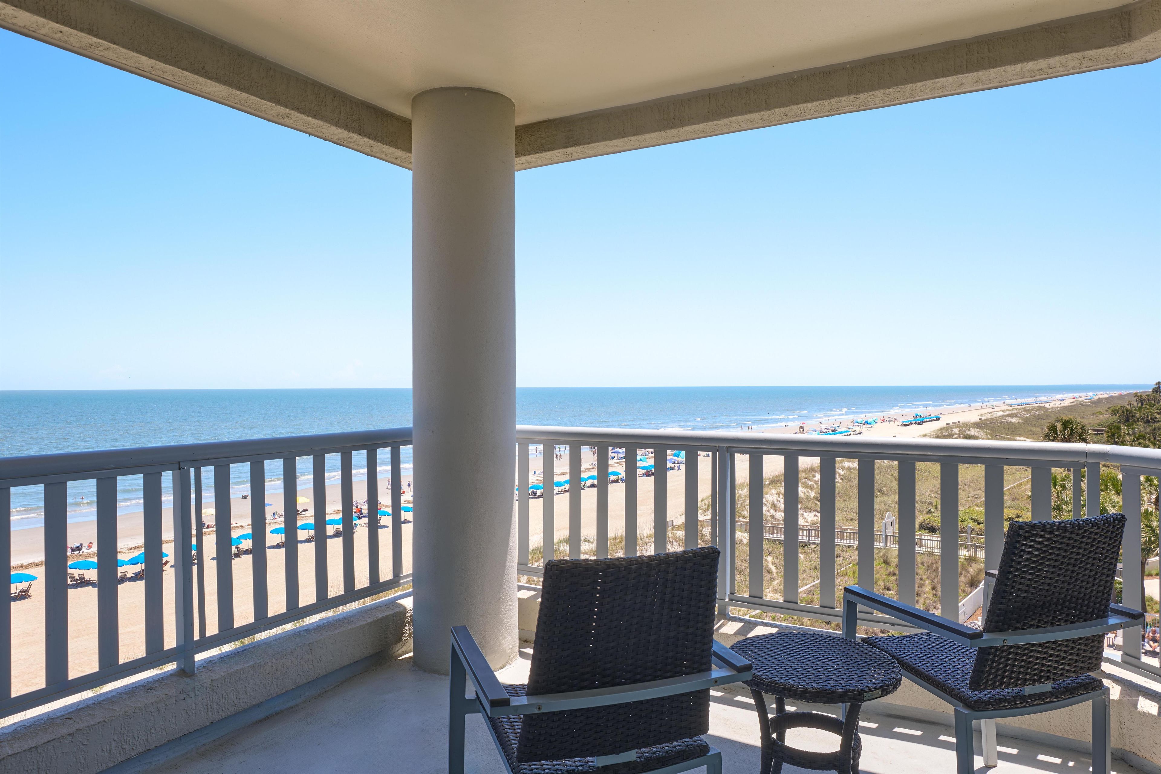A balcony with two wicker chairs, a white railing, and a view of the beach and ocean.