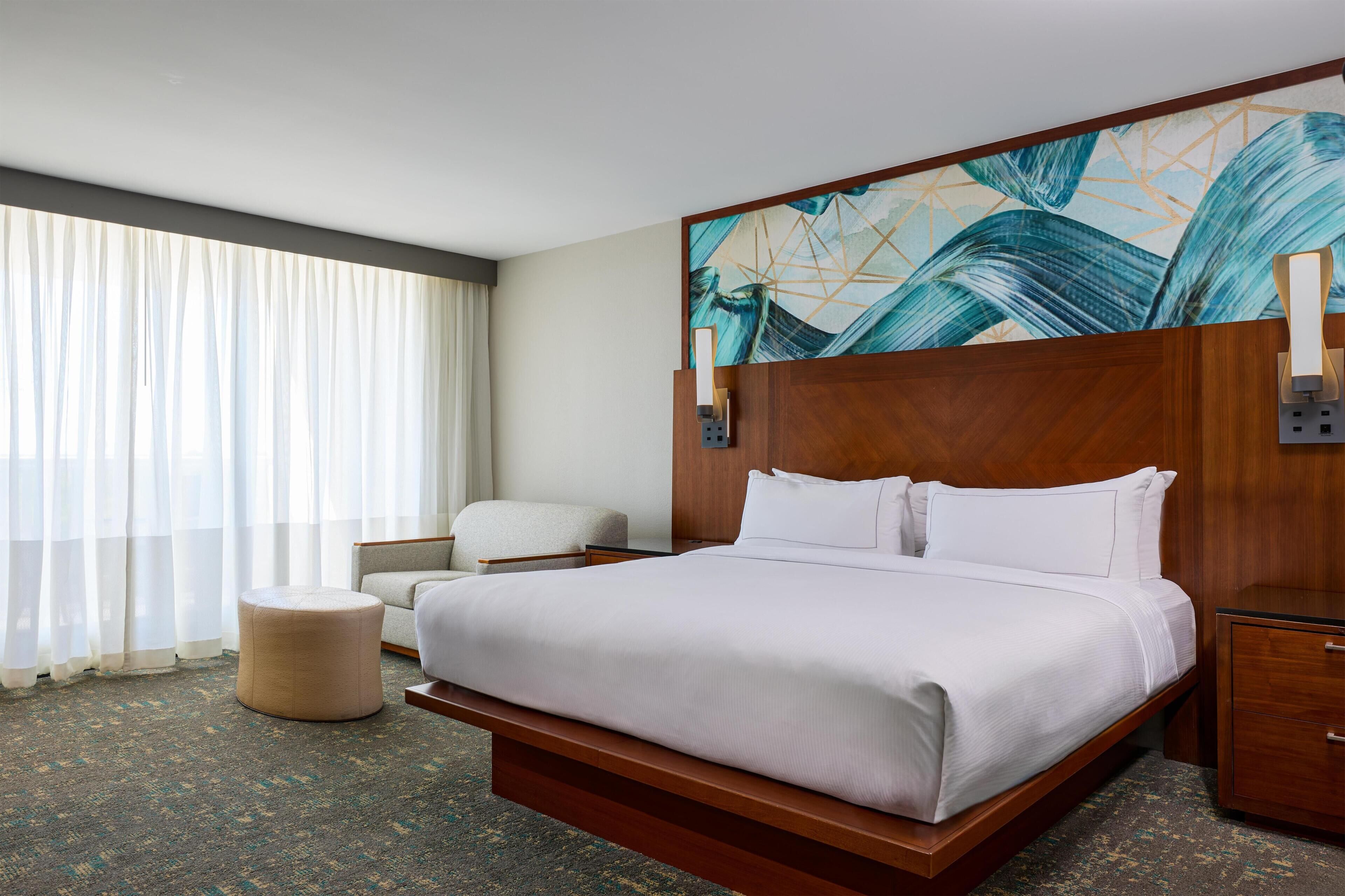 A hotel room with a large bed, a wooden headboard, a nightstand, a chair, and a painting on the wall.