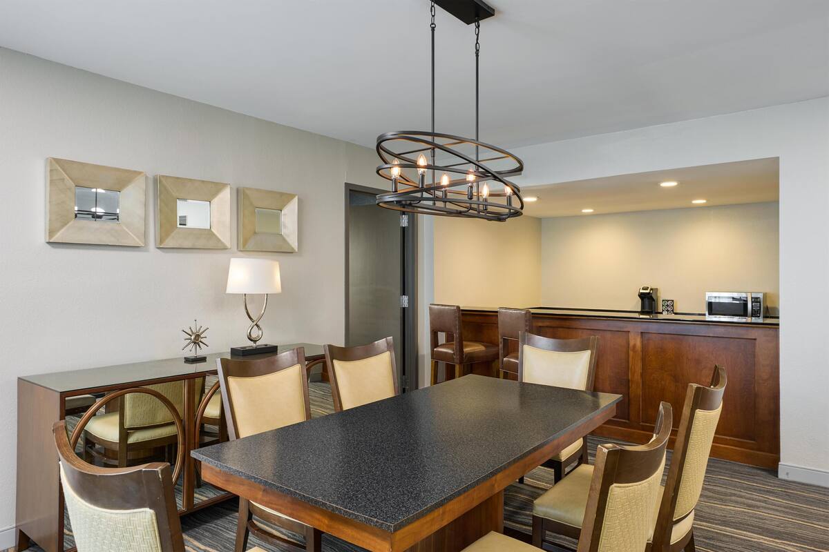 A hotel dining area with a rectangular table, six chairs, a chandelier, a microwave, and a small decorative item on the wall.