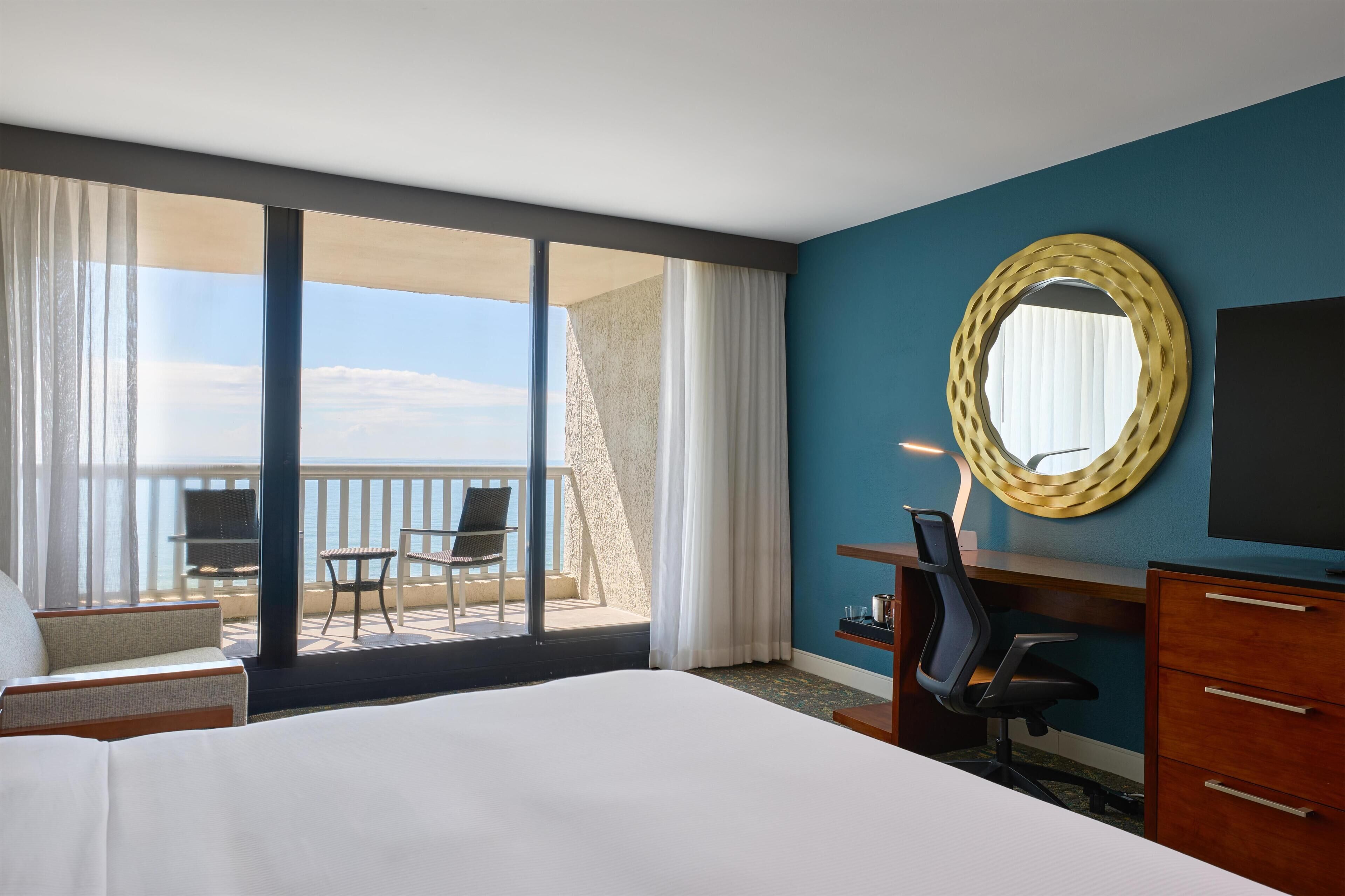 A hotel room with a large bed, a desk, a chair, a TV, and a balcony with a view of the sea.
