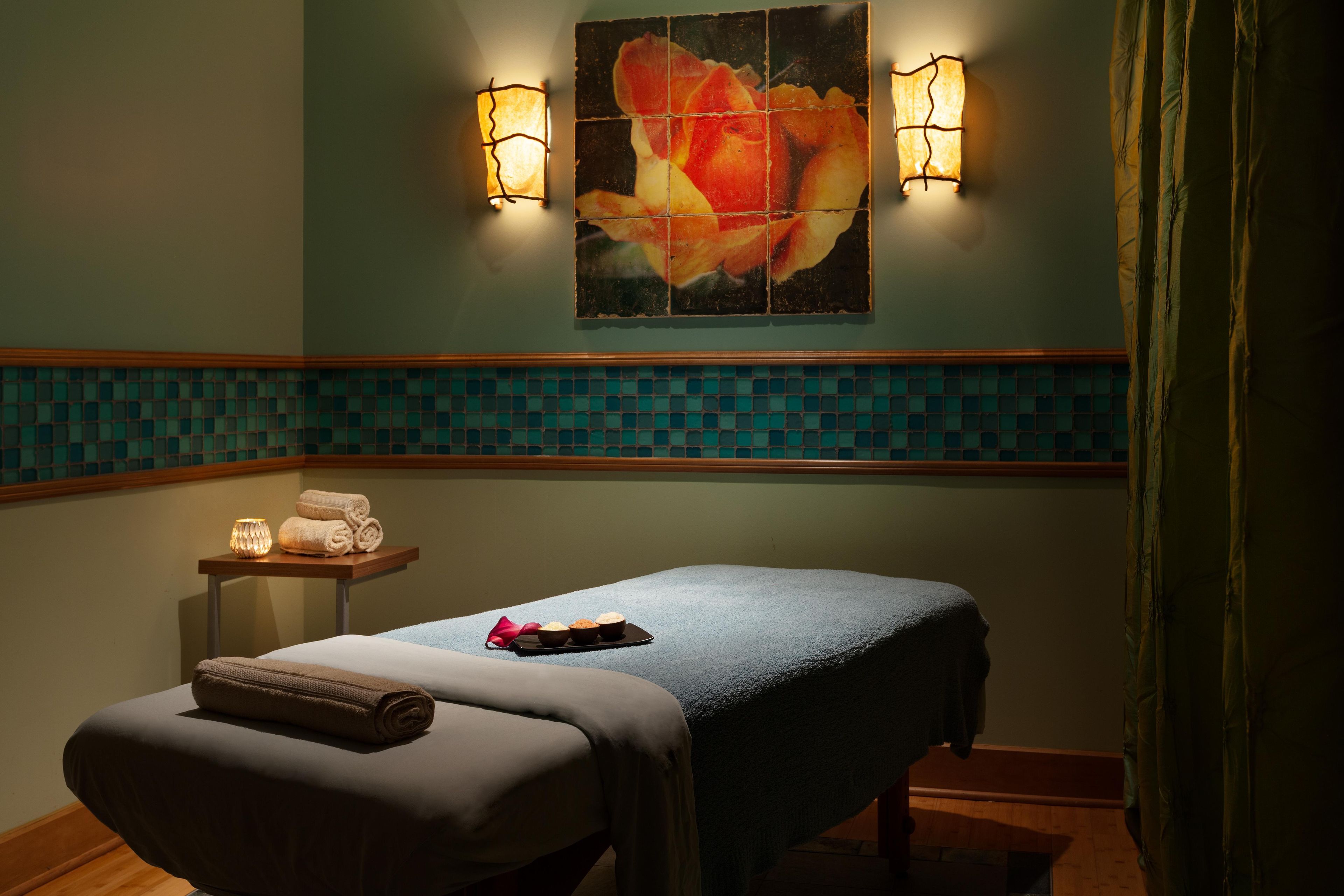 A spa room with a massage table, towels, a small table with candles, and a wall-mounted painting.