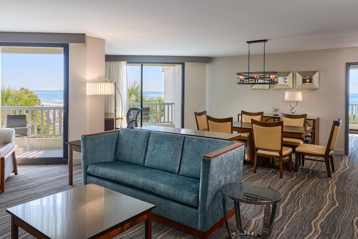 A hotel room with a balcony, a sofa, a dining table with chairs, and a view of the ocean.