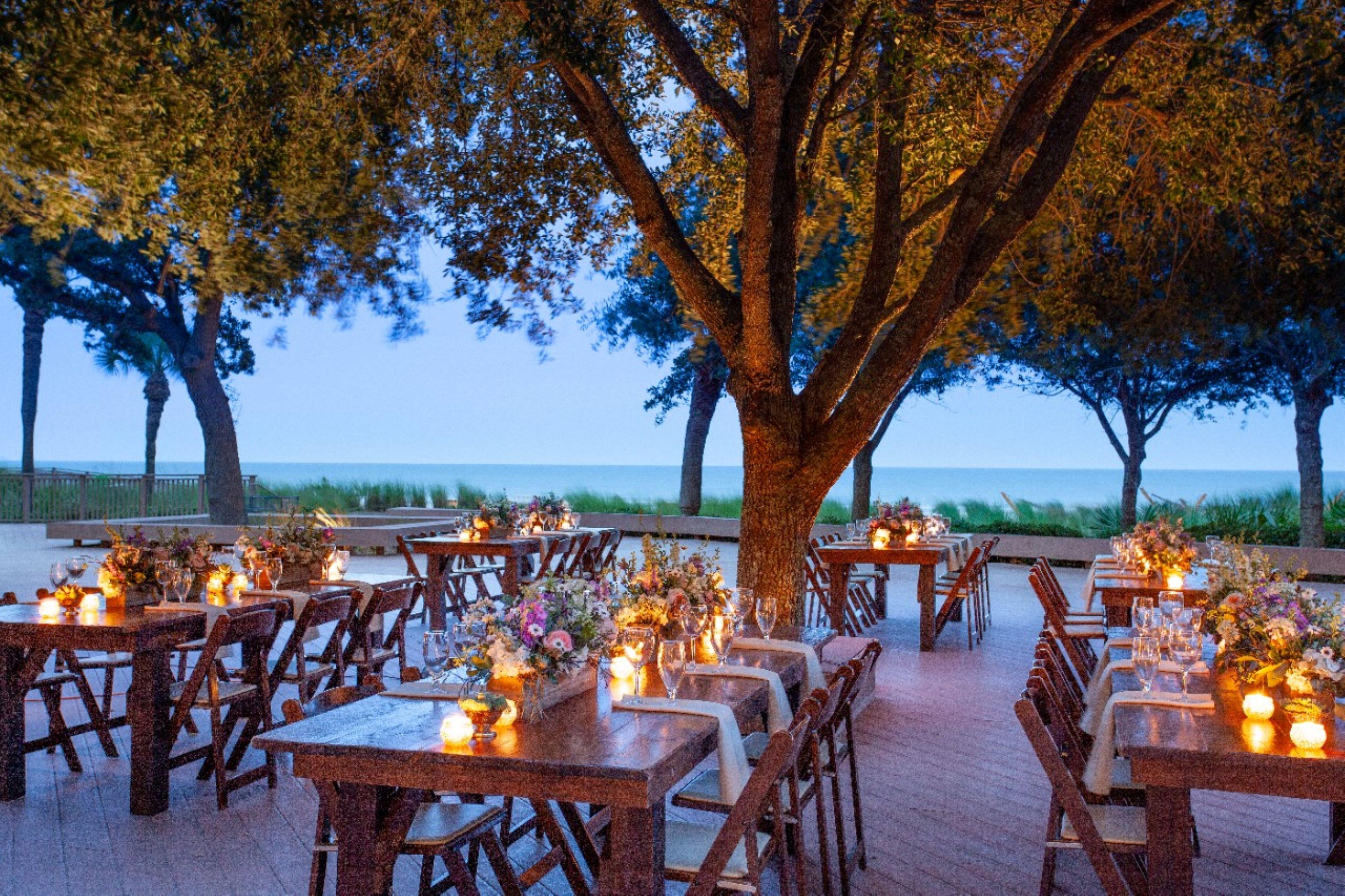 An outdoor dining area with tables set for guests, surrounded by trees and overlooking a body of water.