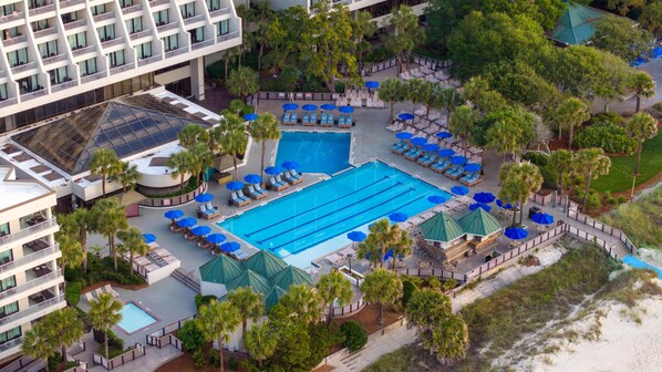 An aerial view of a hotel with a central swimming pool, multiple lounge areas with umbrellas, and surrounding greenery.