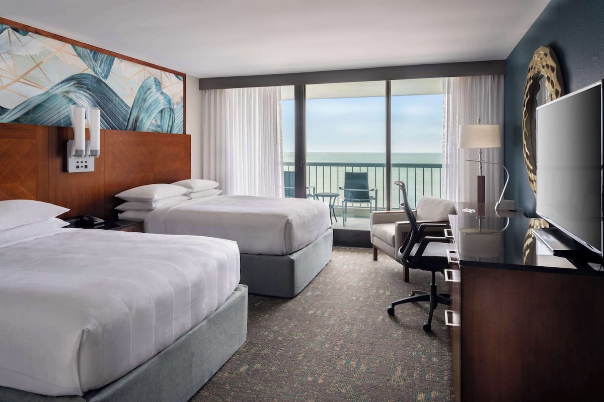 A hotel room with two beds, a desk, a chair, a TV, and a balcony with a view of the ocean.