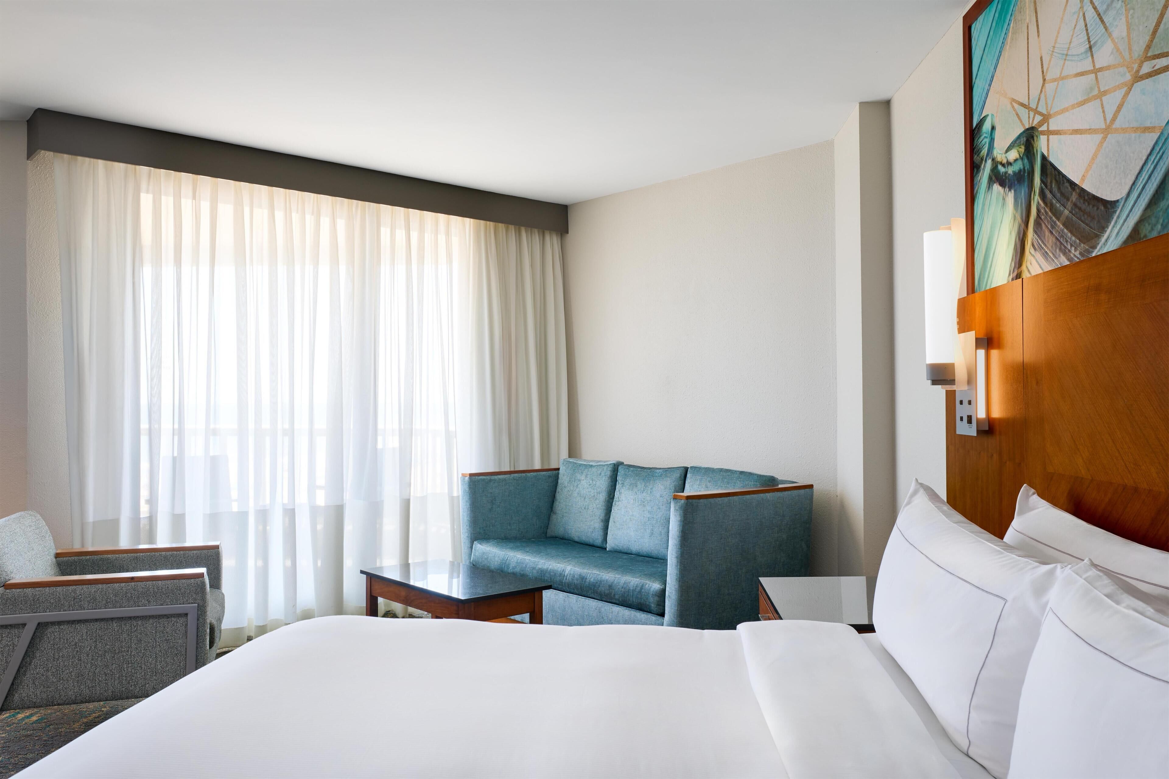 A hotel room with a large bed, a blue armchair, a small table, and a painting on the wall.