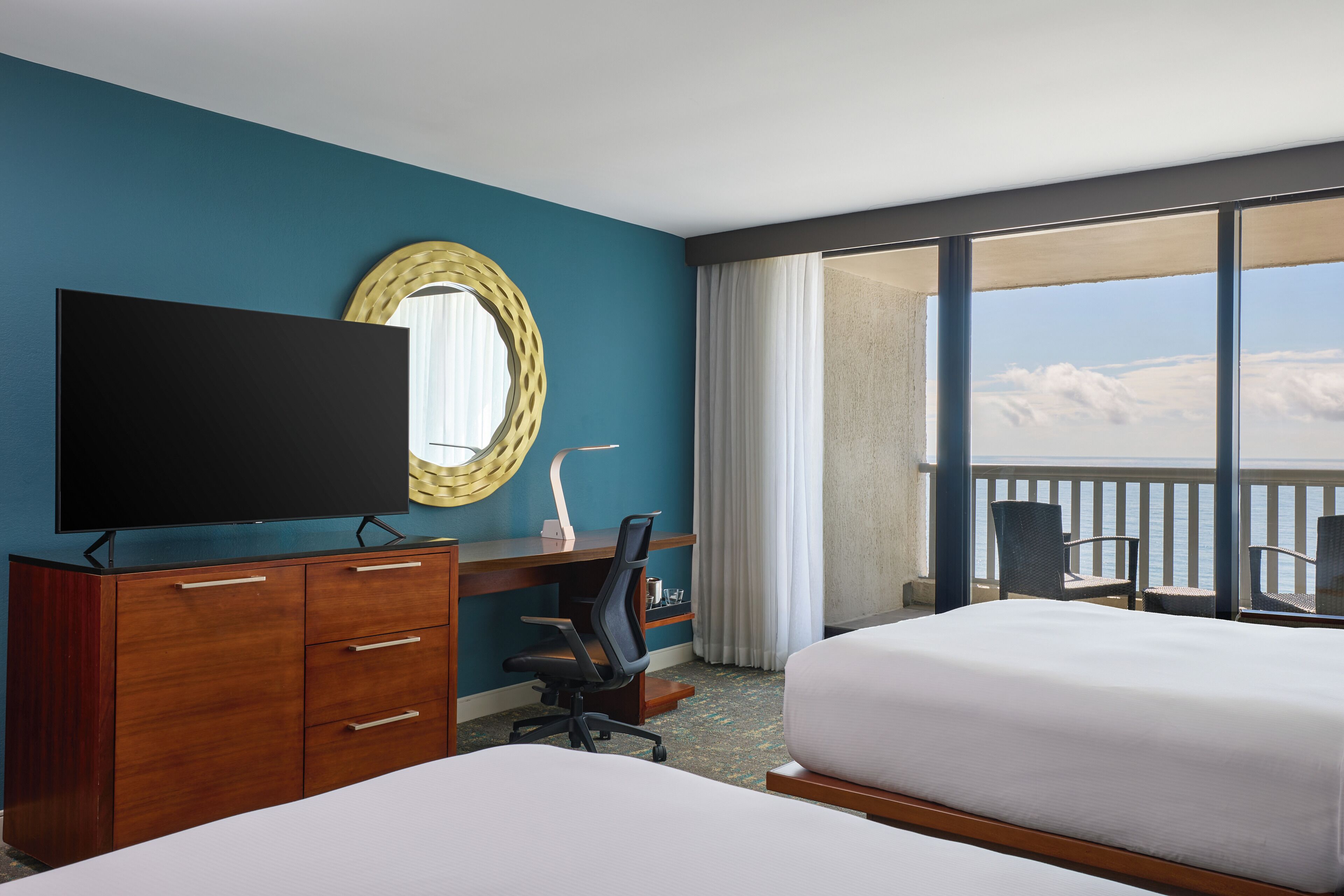 Een hotelkamer met een bed, een bureau met een televisie, een stoel, een groot raam met uitzicht op de wolken en een balkon.