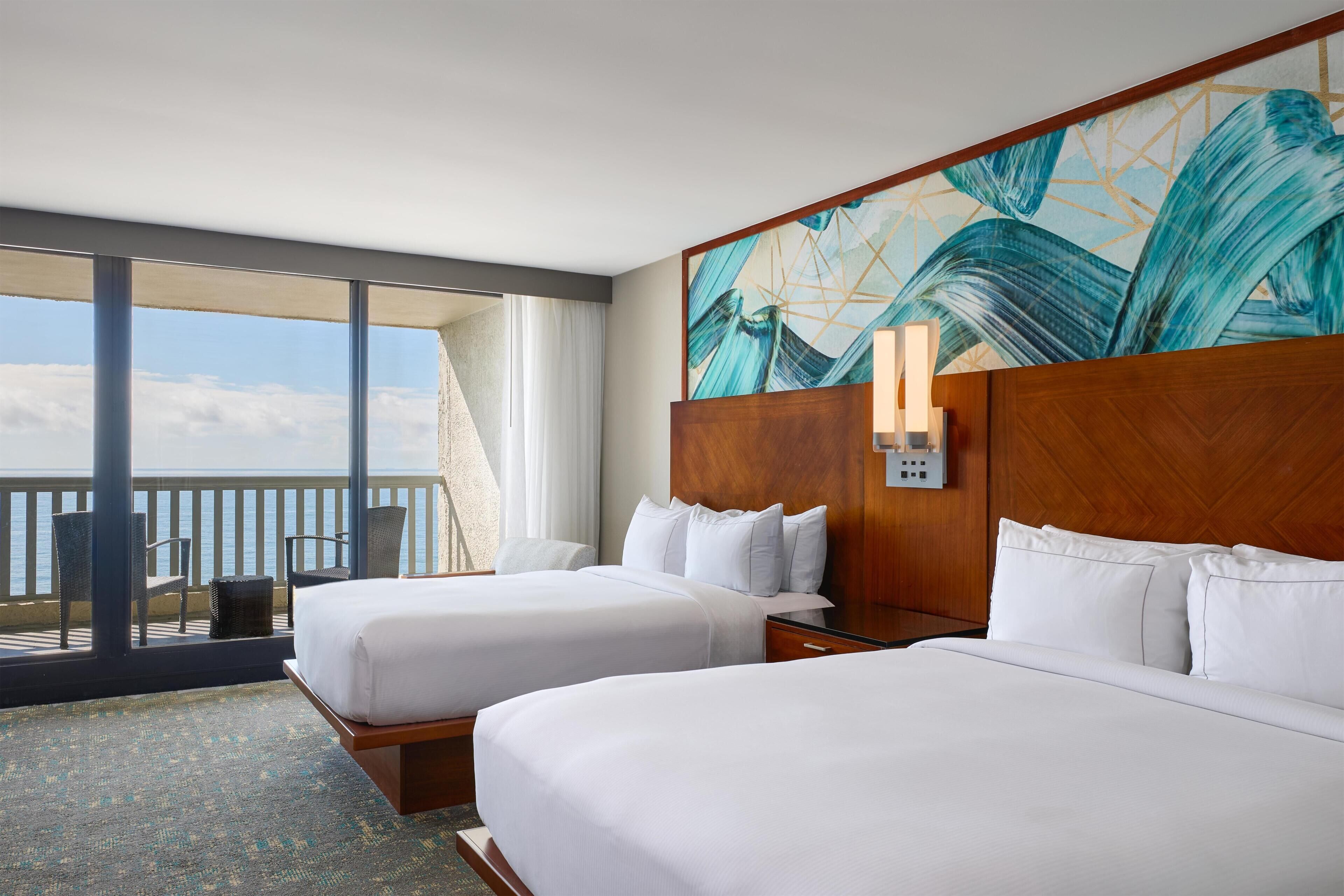 A hotel room with two beds, a large window with a view of the sea, and a painting on the wall.