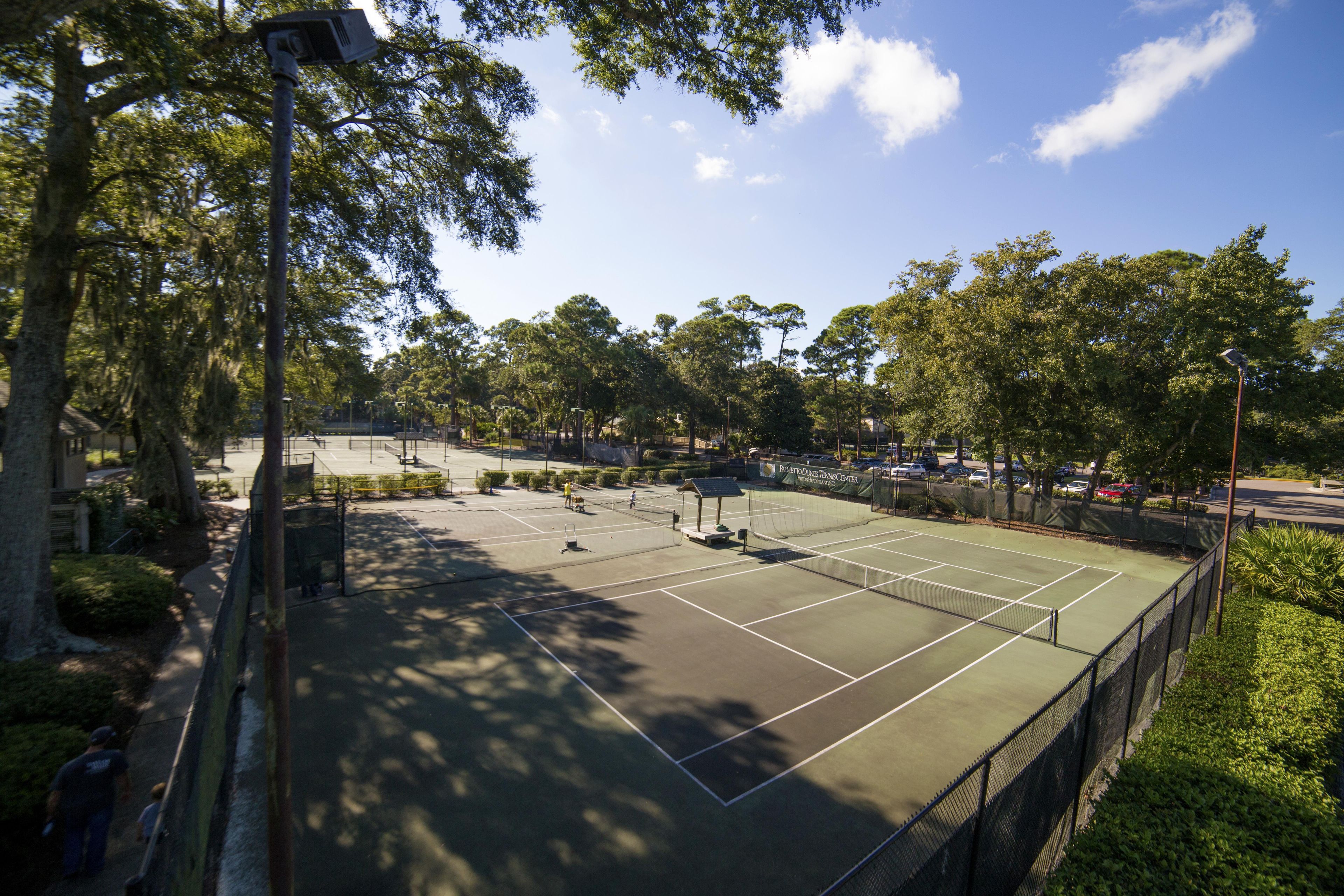 Un court de tennis en plein air, doté d’un filet, entouré d’arbres et délimité par une clôture.