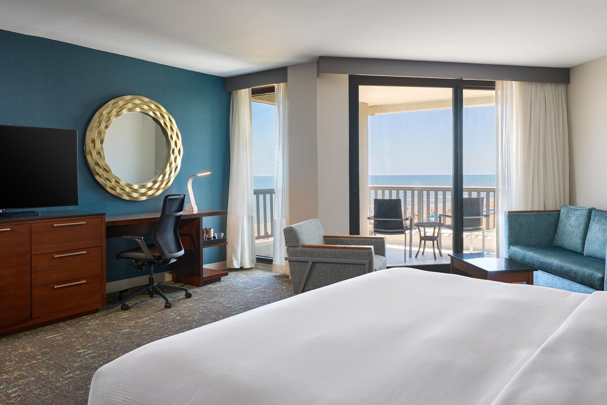 A hotel room with a bed, desk, chair, and a view of the beach.