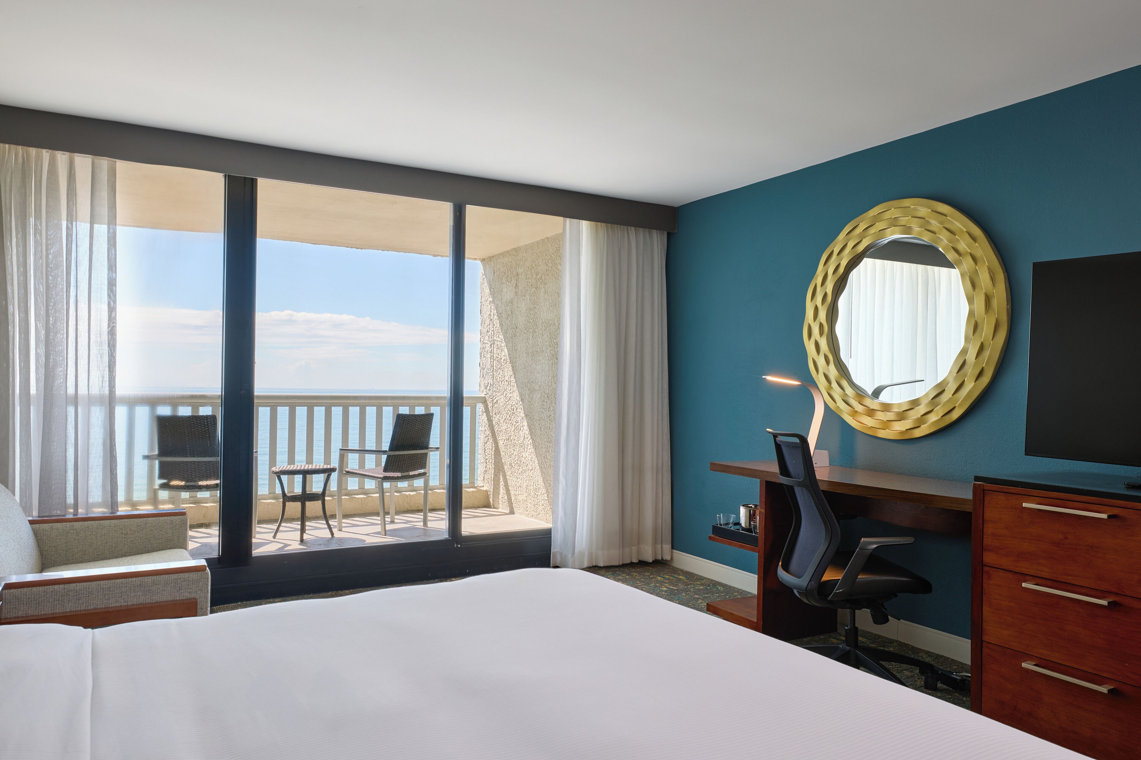 Une chambre d’hôtel avec un grand lit, un bureau, une chaise, une télévision et un balcon avec vue sur l’océan.