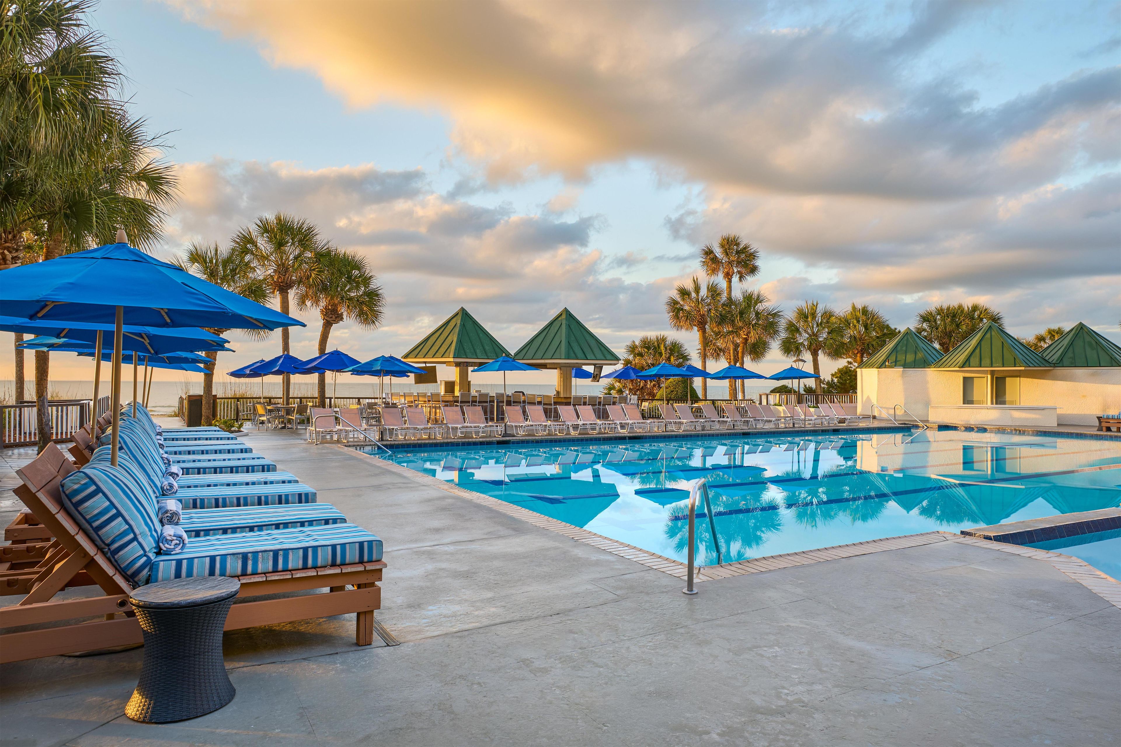 A poolside area with lounge chairs, umbrellas, and palm trees.