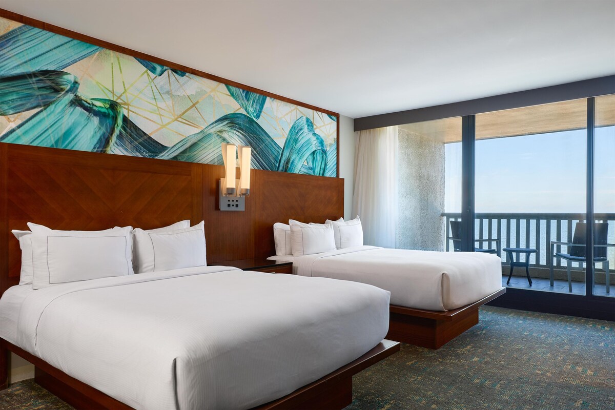 A hotel room with two beds, a large abstract painting, a nightstand, and a balcony with chairs.