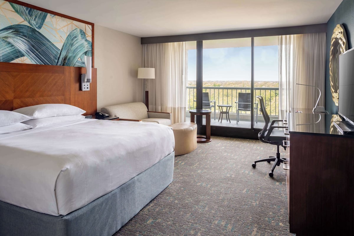 A hotel room with a large bed, a desk, a chair, a lamp, and a view of the outdoors.