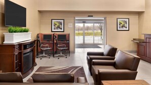 A hotel lobby with a flat-screen TV, a fireplace, leather sofas, a desk with two monitors, and a large glass door leading outside.