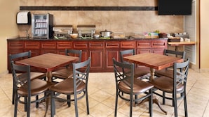 A kitchen with a buffet setup, including a refrigerator, a microwave, and a coffee machine. There are two wooden tables with chairs, and a television mounted on the wall.