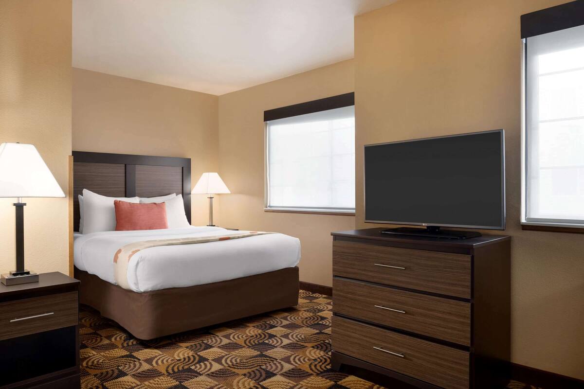 Ein Hotelzimmer mit Bett, zwei Nachttischlampen, einem Flachbildfernseher, einem Schminktisch und einem Fenster mit Jalousien.