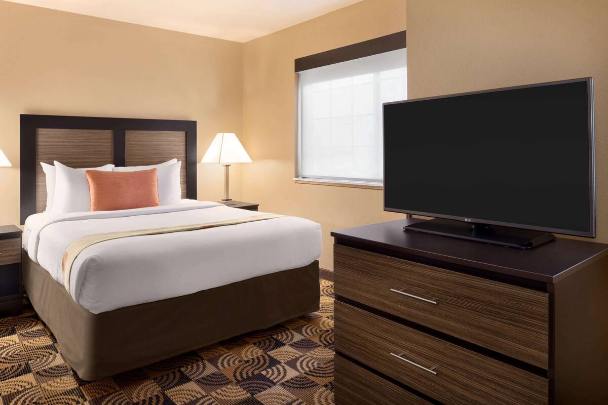 Ein Hotelzimmer mit Bett, Fernseher auf einer Kommode, Nachttisch mit Lampe und einem Fenster mit Jalousien.