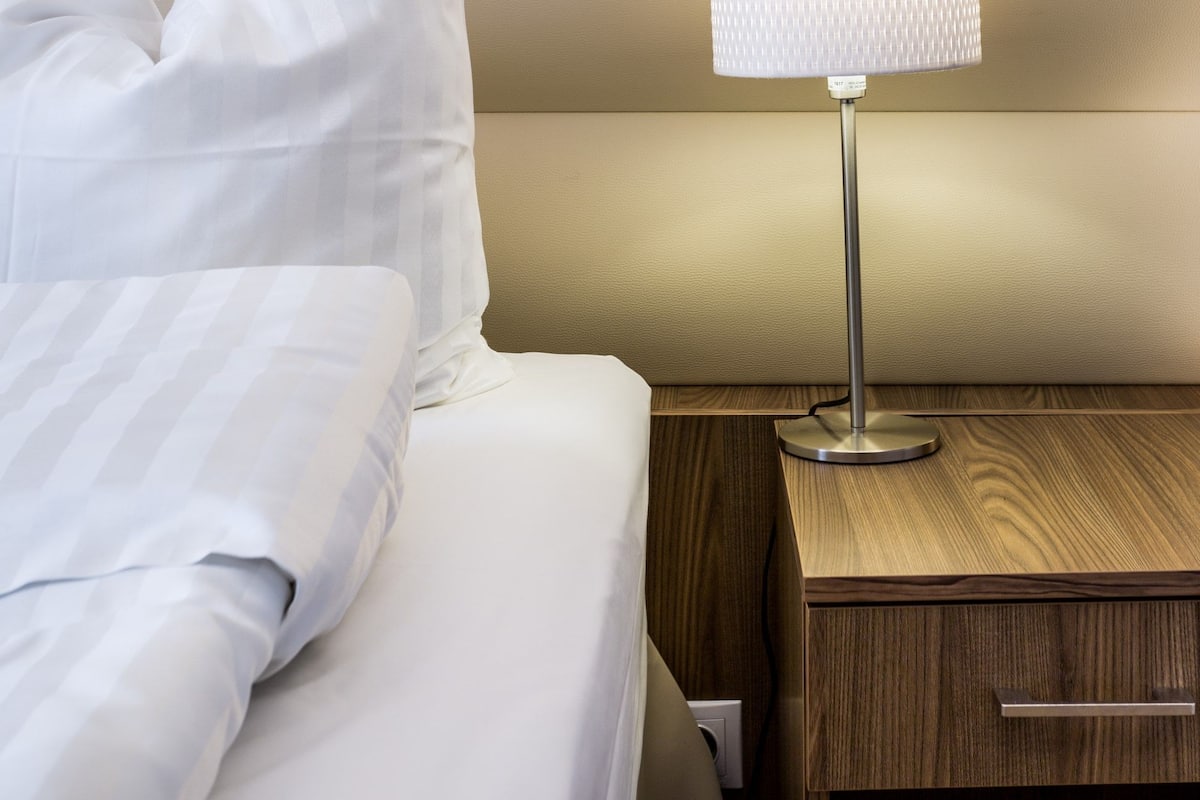 A neatly made bed with white linens, a bedside lamp with a white shade, and a wooden nightstand with a drawer.