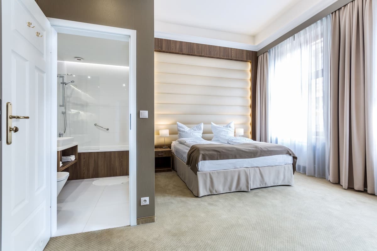 A hotel room with a large bed, a walk-in closet, a shower, and a view of the city through sheer curtains.