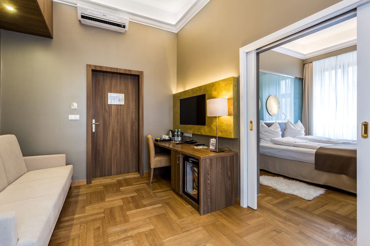 A hotel room with a wooden herringbone floor, a sofa, a desk with a flat-screen TV, a minibar, and a bed with a large window.