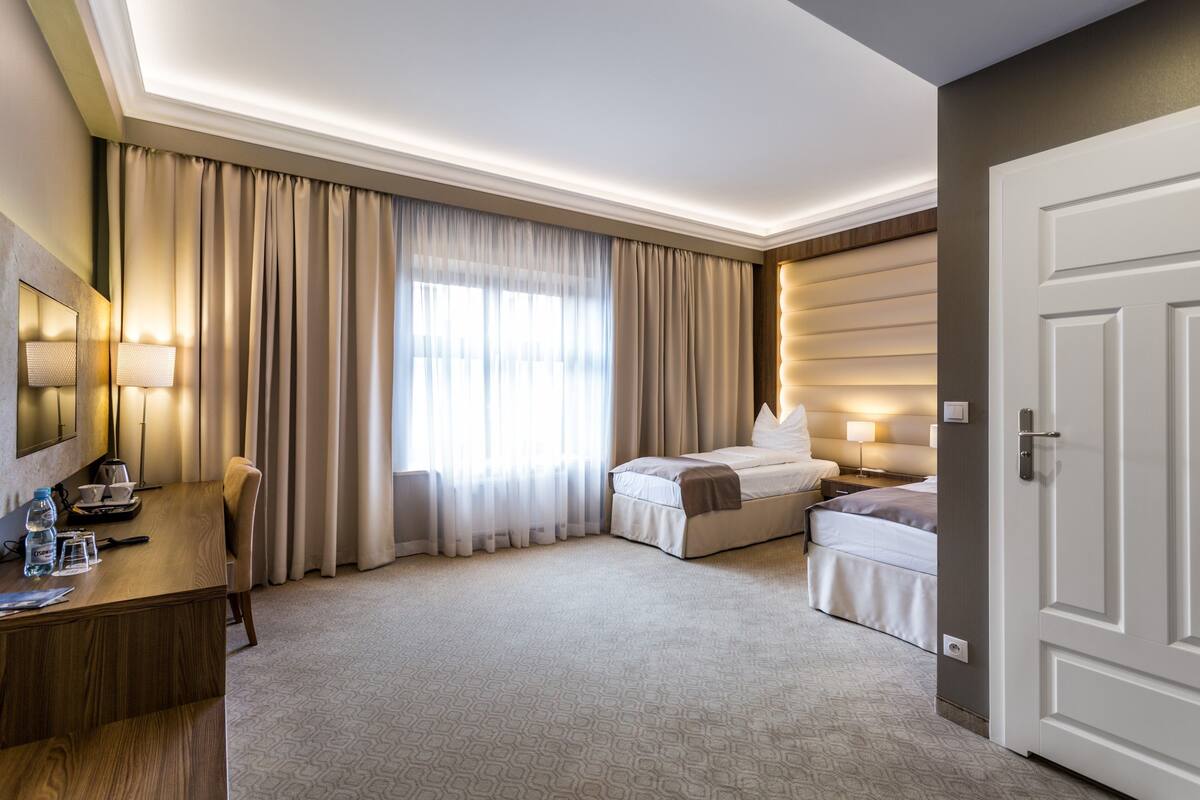 A hotel room with a desk, two beds, a large window with curtains, and a door.