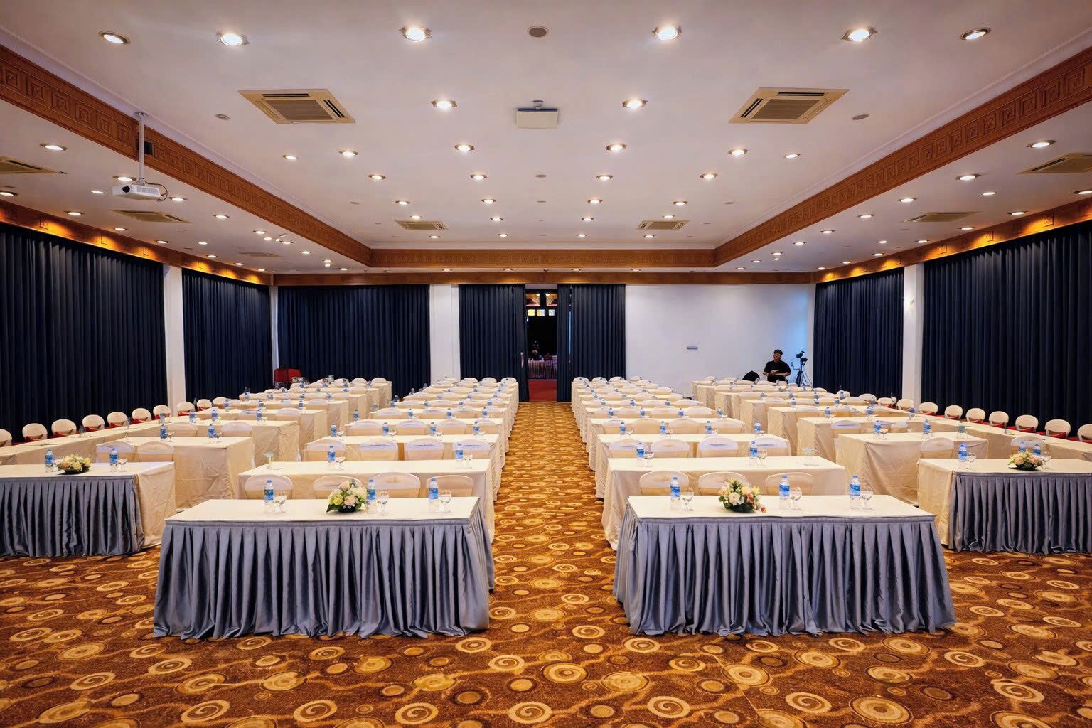 A conference room with rows of tables and chairs, equipped with water bottles and floral centerpieces.