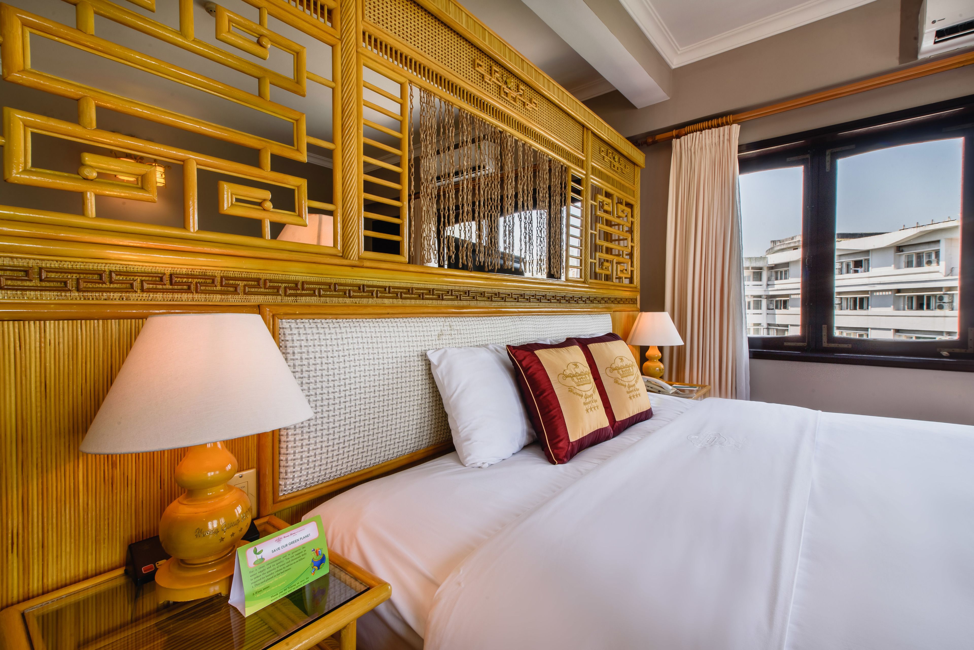 A hotel room with a large bed, two bedside lamps, a decorative headboard, and a view of buildings through the window.
