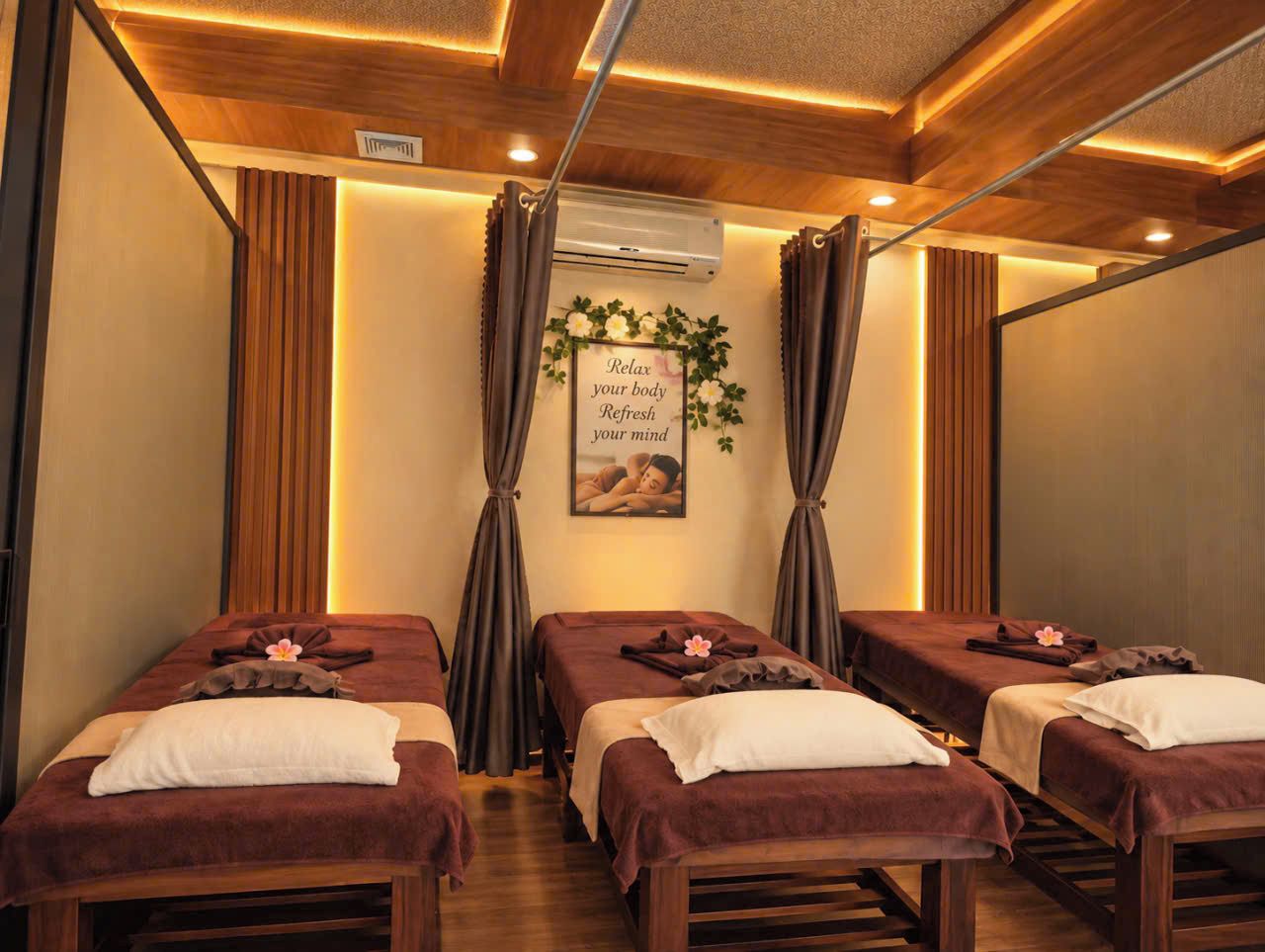 Un cuarto de spa con tres camillas de masaje, cada una con una toalla y una flor. Hay un aire acondicionado de pared y un cartel con un mensaje de relajación.
