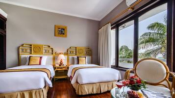 A hotel room with two beds, a dining table with fruit and drinks, and a view of palm trees through large windows.