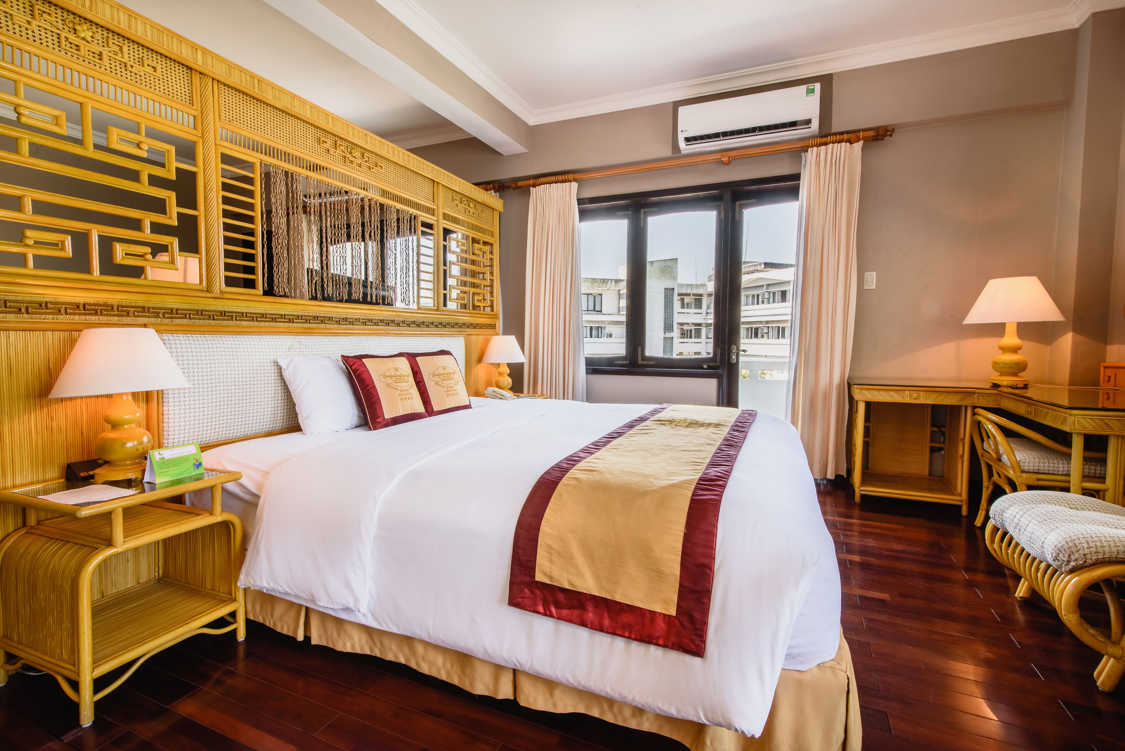 A hotel room with a large bed, two bedside tables with lamps, a decorative headboard, a window with curtains, and a view of buildings outside.