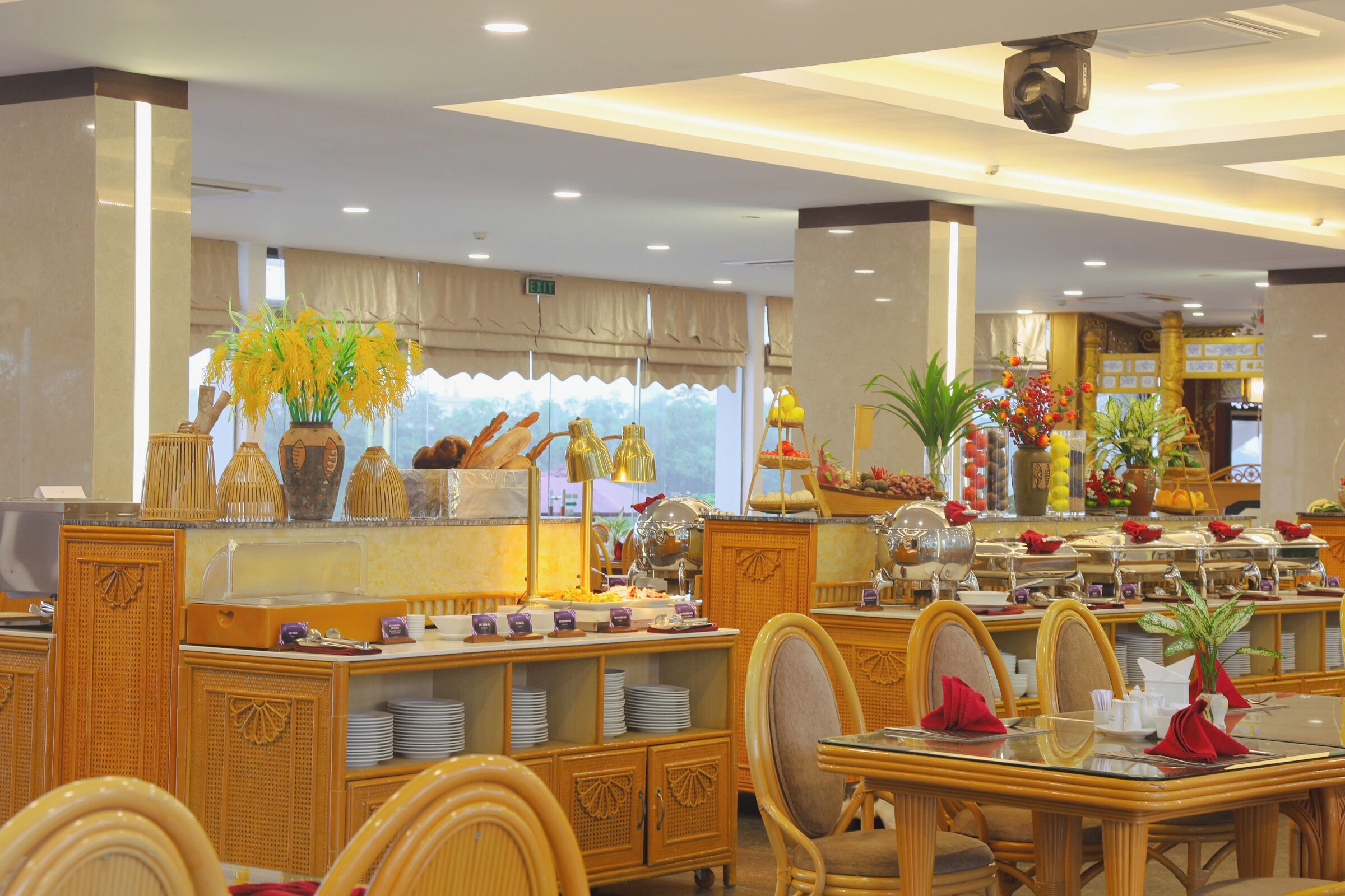 Un comedor de hotel con mobiliario de mimbre, un mostrador de buffet con diversas opciones de comida y plantas decorativas.
