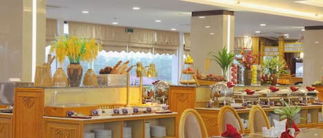 Un comedor de hotel con mobiliario de mimbre, un mostrador de buffet con diversas opciones de comida y plantas decorativas.