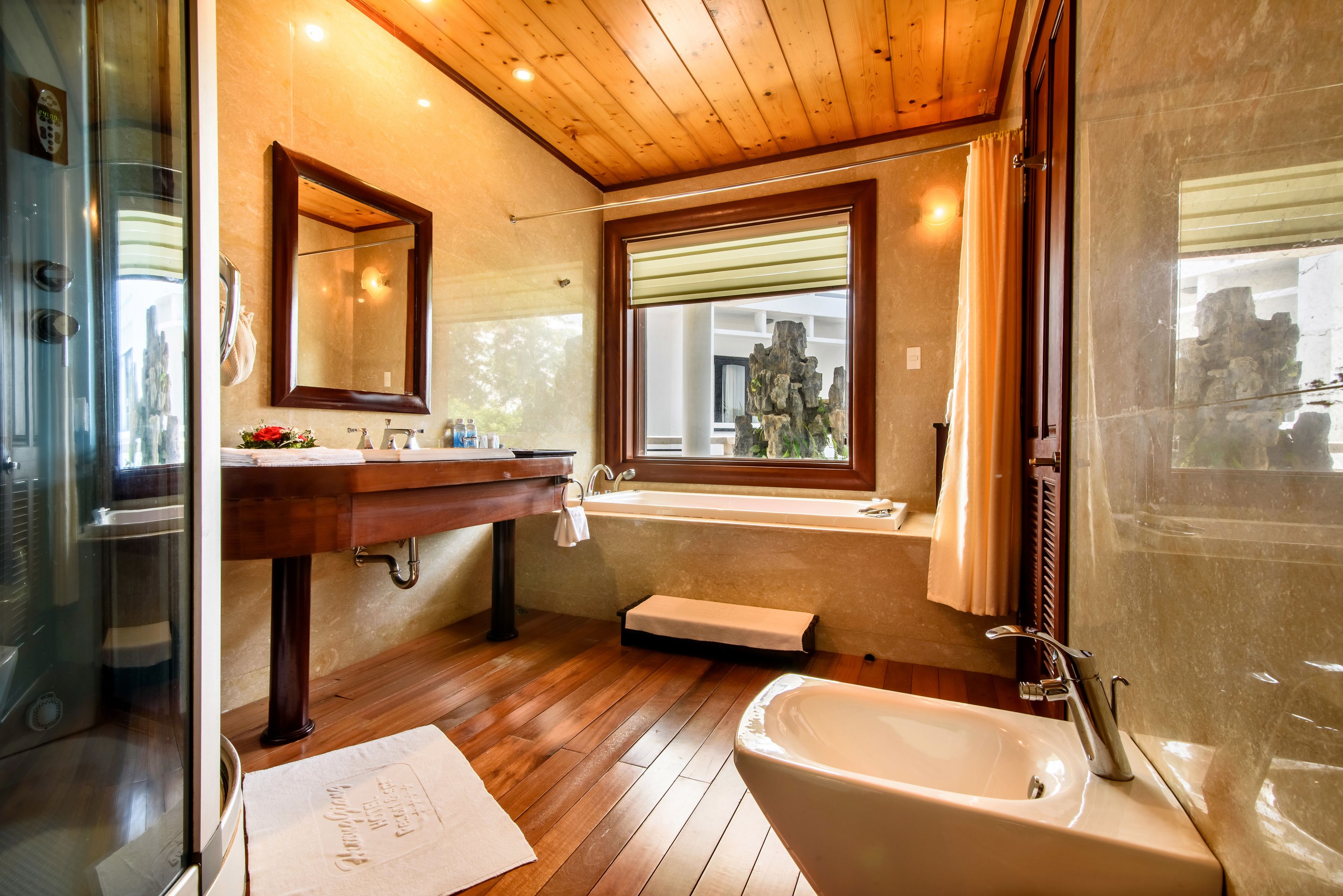 A bathroom with a wooden floor, a sink with a mirror, a bathtub, and a window with a view of a rocky landscape.