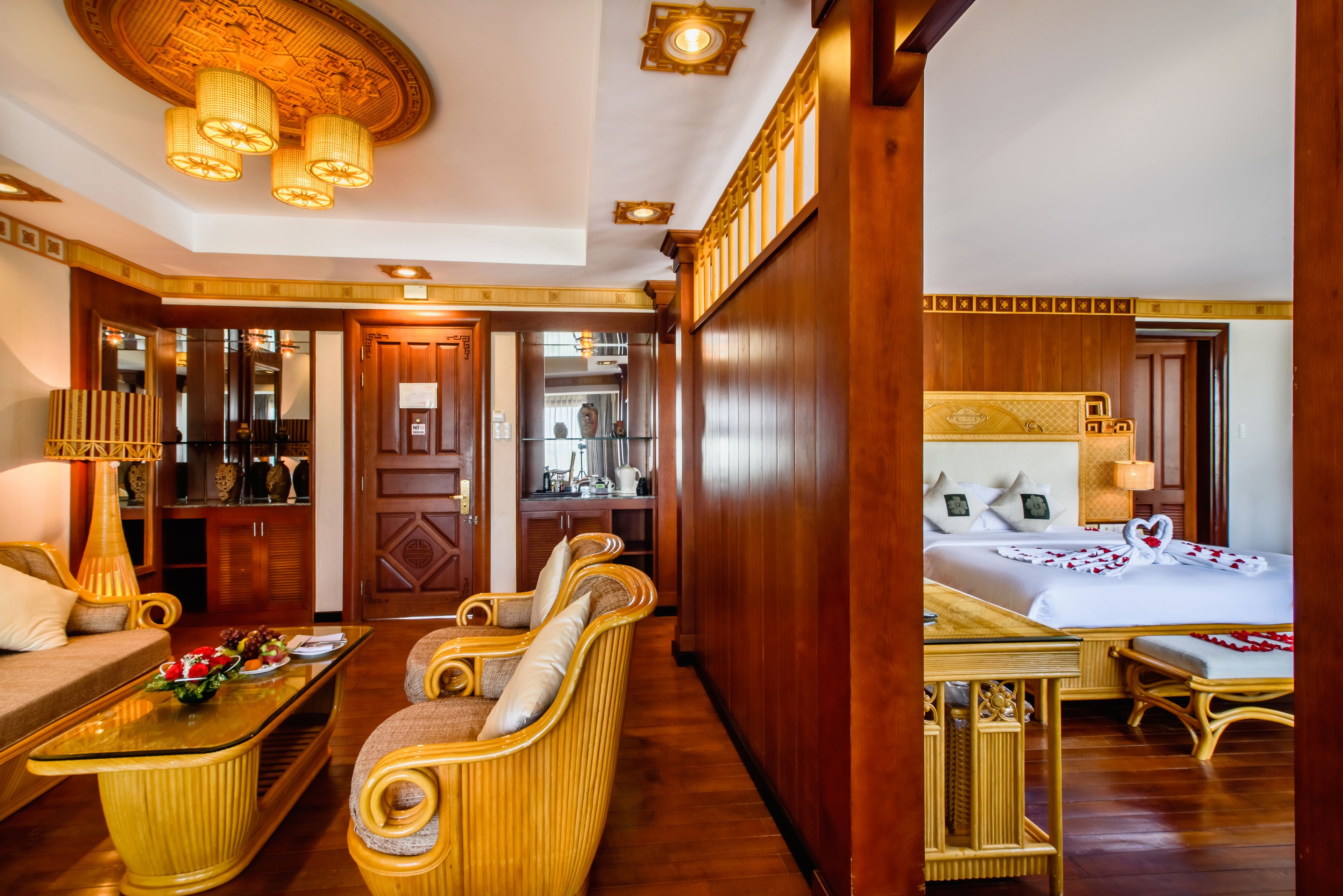 A hotel room with a wooden floor, a bed with a white bedspread and a heart-shaped decoration, a wooden wardrobe, a sitting area with wicker furniture, and a dining table with a vase of flowers.
