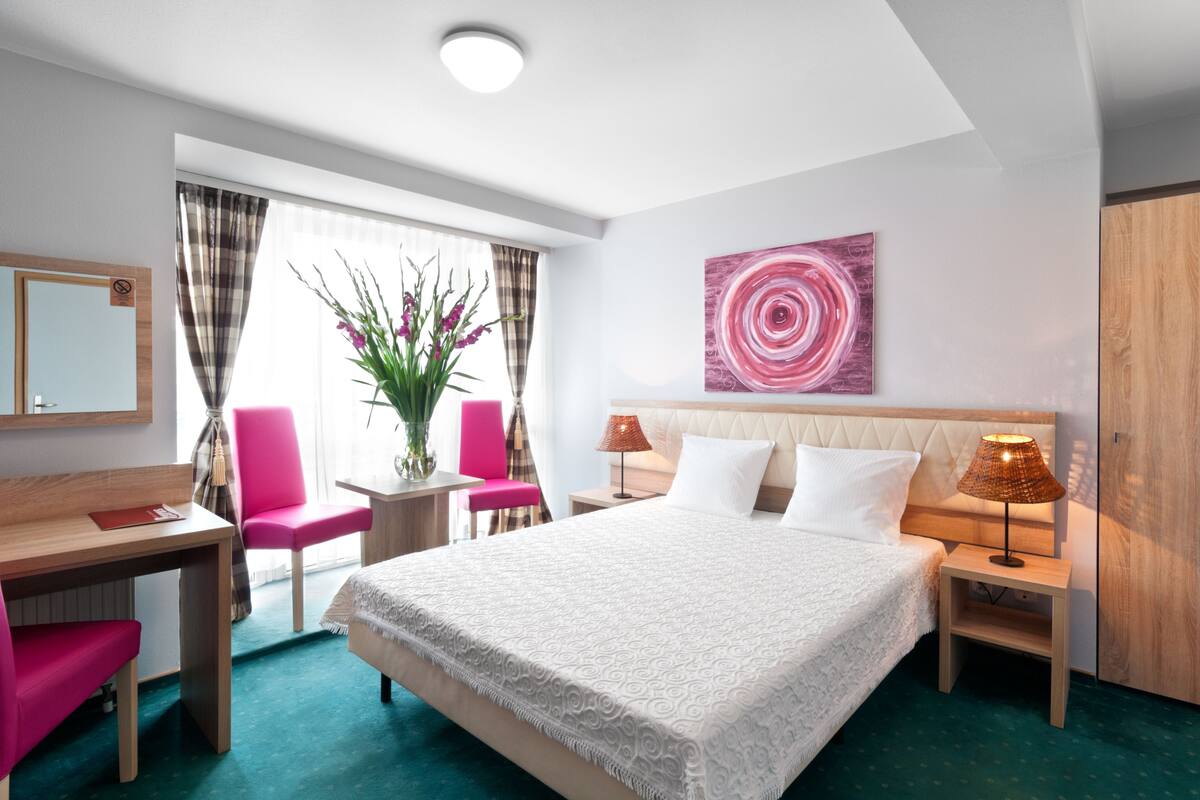 A hotel room with a large bed, a desk, a chair, a nightstand, and a window with curtains.