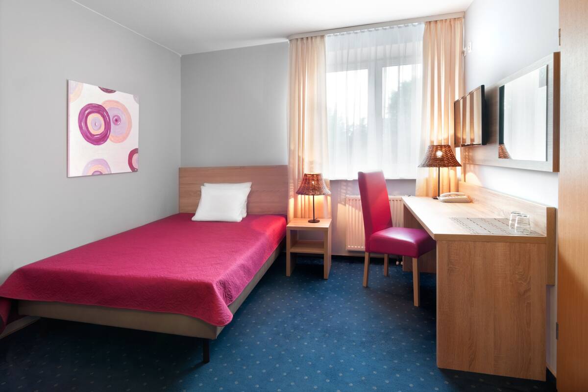 A hotel room with a single bed, a desk, a chair, and a window with curtains.