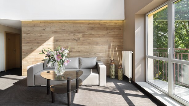 A modern living room with a wooden accent wall, a sofa, a glass coffee table, and a large window offering a view of greenery. - Petrus Hotel (Kraków)