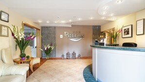 A reception area with a reception desk, a clock, framed certificates, and a floral arrangement. - Petrus Hotel (Kraków)
