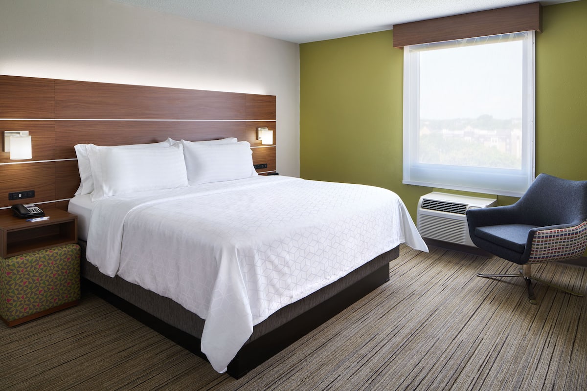 A hotel room with a large bed, bedside tables, a chair, a window, and an air conditioning unit.