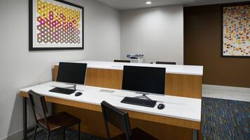 A conference room with two desks, each with a computer monitor, keyboard, and mouse. There are two chairs at each desk. The room has a whiteboard, a printer, and two framed artworks on the walls.