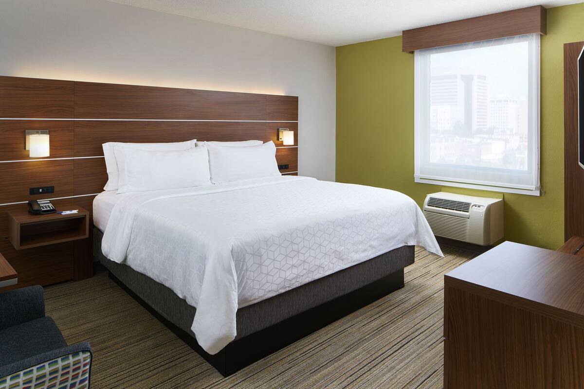 A hotel room with a large bed, a nightstand with a phone, a desk, and a window with a view of buildings.