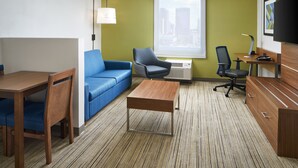 A hotel room with a wooden desk, blue sofa, and a small table. There is a window with a view of buildings, a flat-screen TV, and a lamp. - Holiday Inn Express Richmond Downtown by IHG (Richmond)