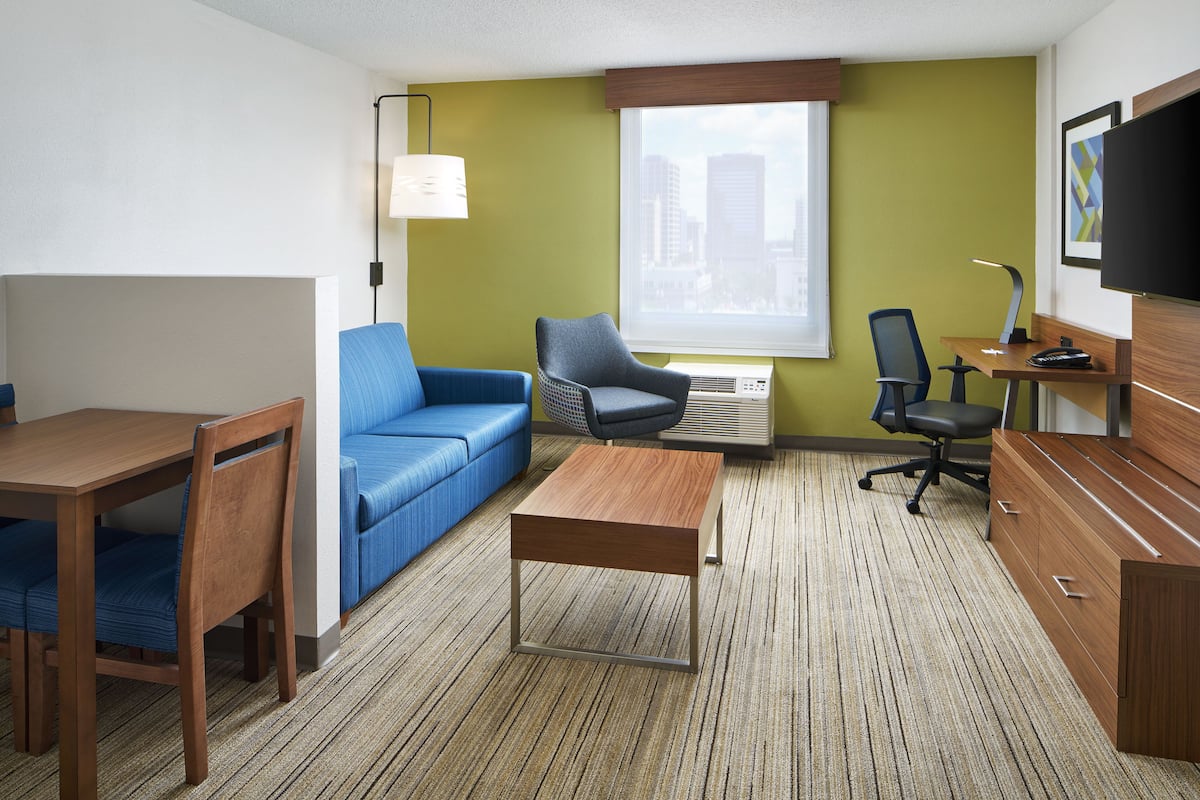 A hotel room with a wooden desk, blue sofa, and a small table. There is a window with a view of buildings, a flat-screen TV, and a lamp.