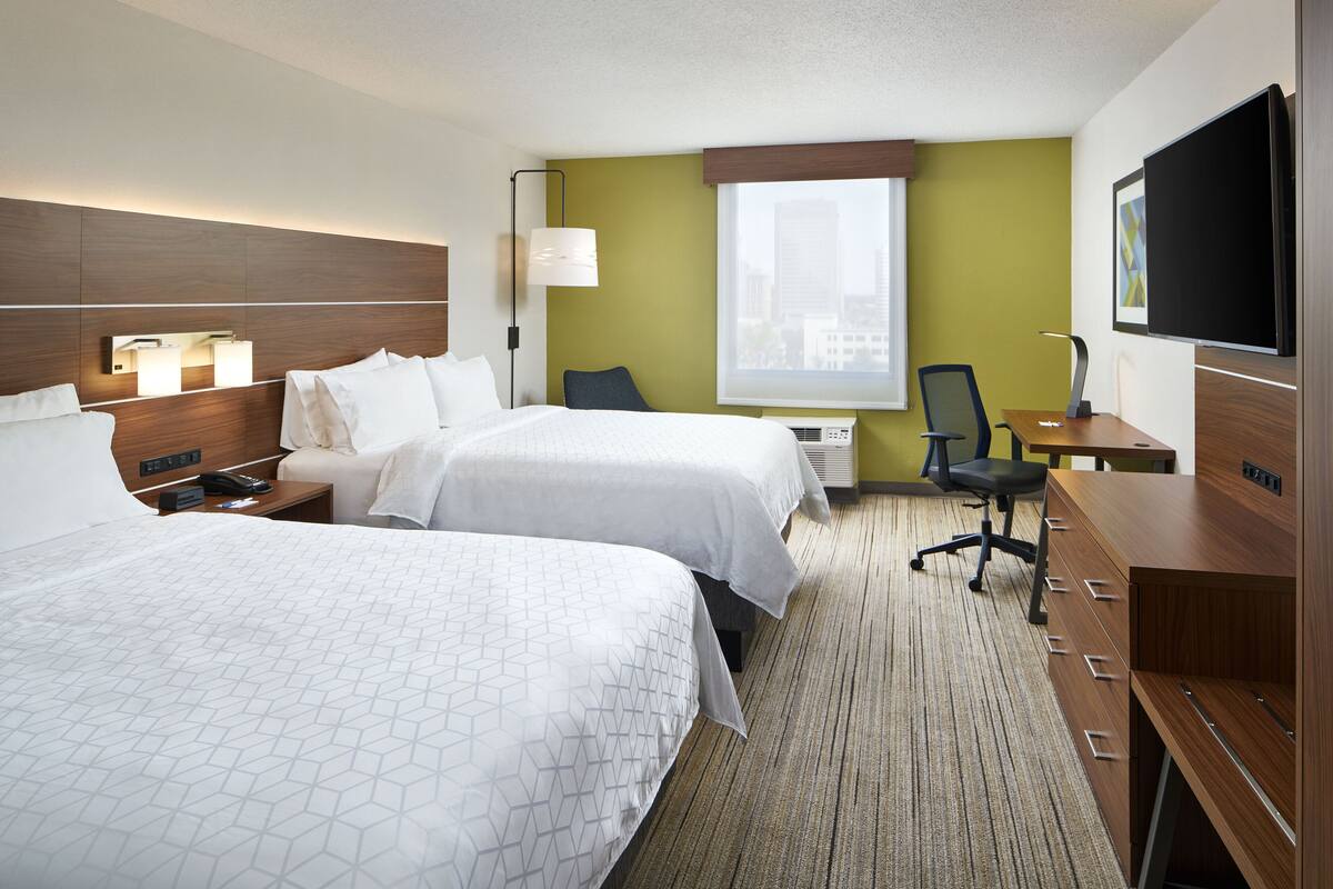A hotel room with two beds, a desk, a chair, a TV, and a window with a city view.