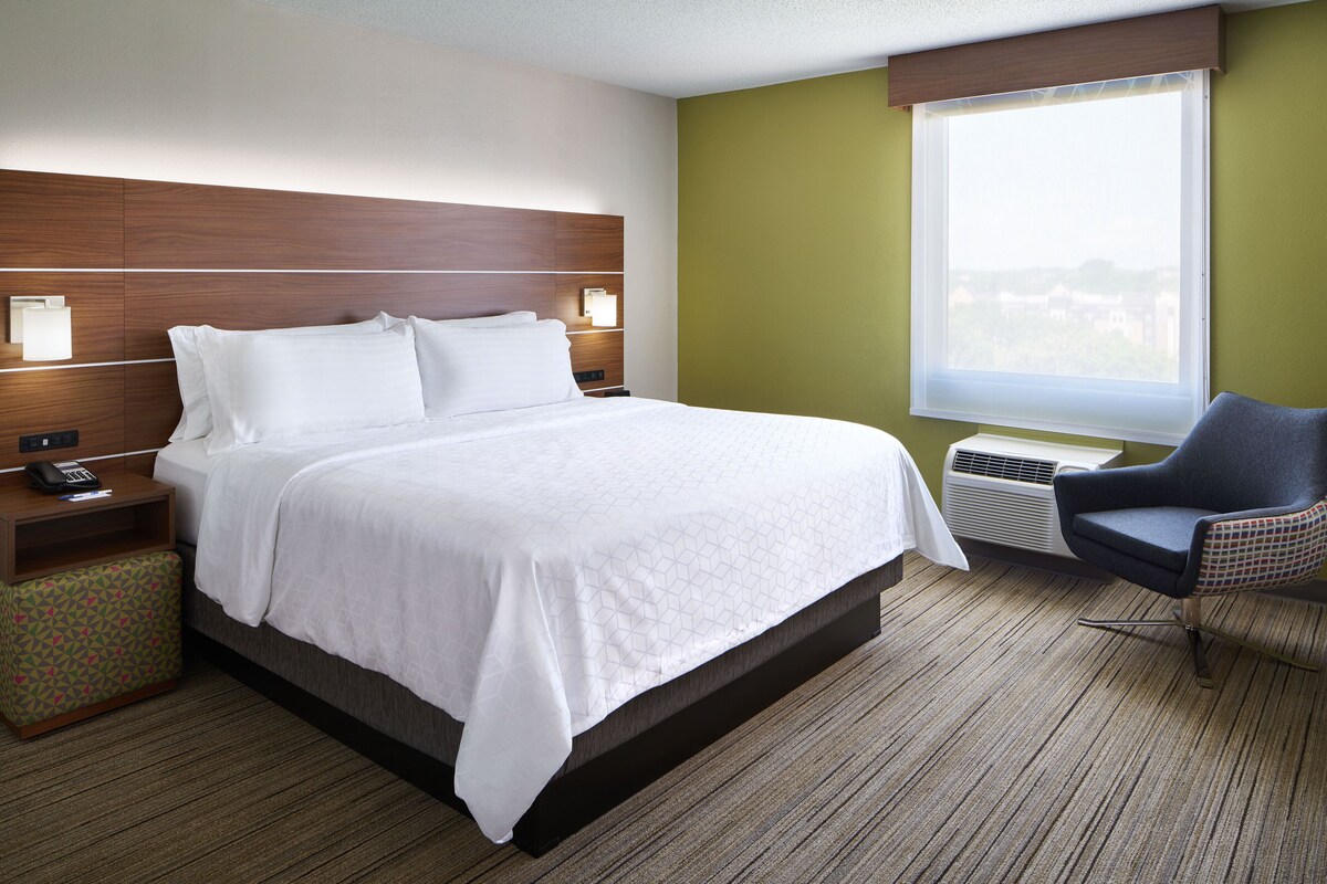 A hotel room with a bed, bedside tables, a chair, a window, and an air conditioning unit.