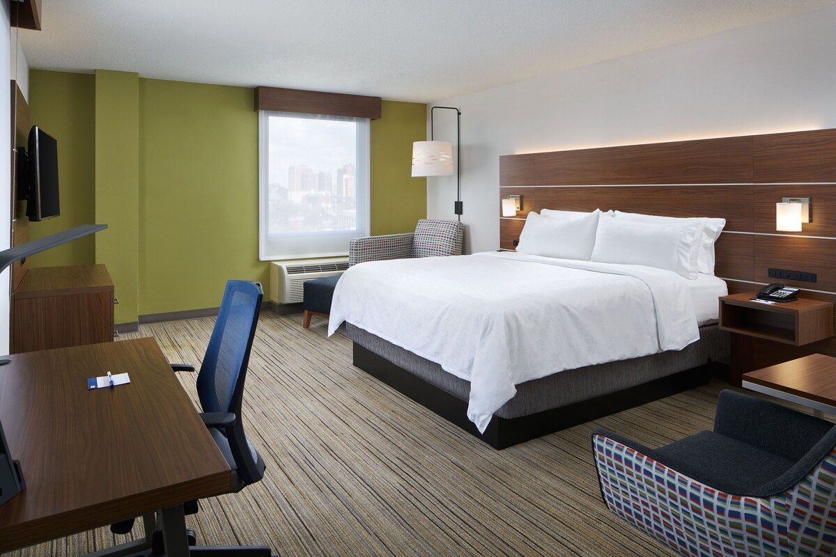 A hotel room with a bed, a desk, a chair, a TV, and a window with a city view.