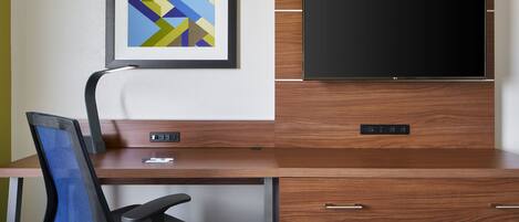 A modern hotel room with a wooden desk, a black office chair, a flat-screen TV mounted on a wooden cabinet, and a framed abstract art piece on the wall.