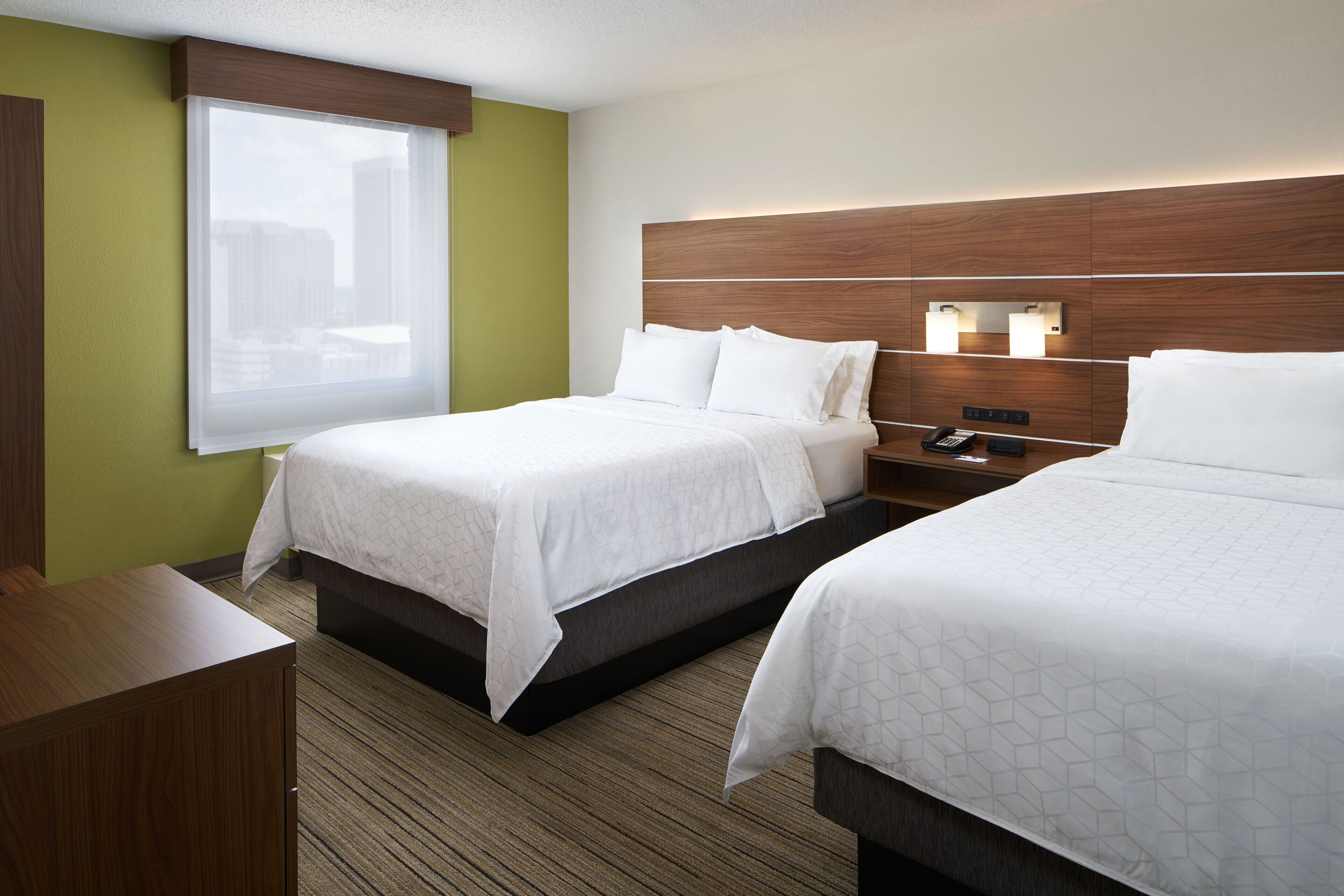 Quarto de hotel com duas camas, uma mesinha de cabeceira, um telefone e uma janela com vista para edifícios.